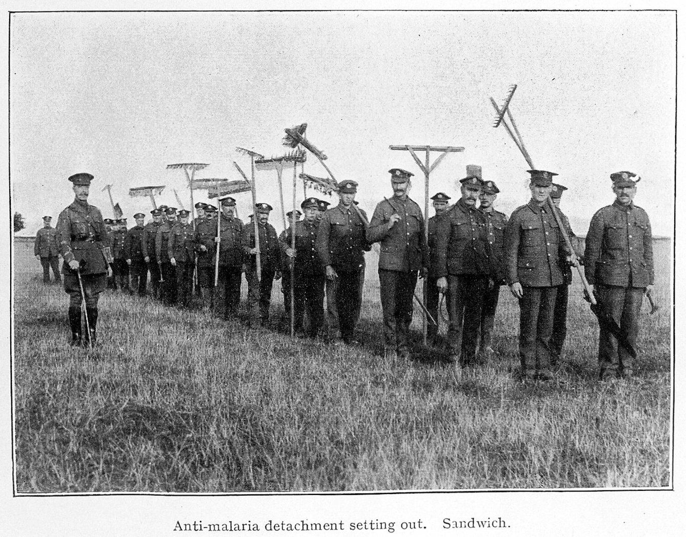 A line of men in military uniform with rakes, spades and other tools over their shoulders standing in a grassy field. Their commander stands to one side of the line. Black and white photograph.