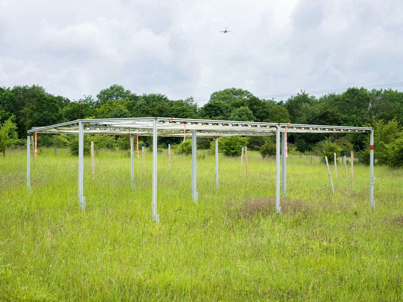 Colour photograph of a wild grassland meadow. In the centre is a large man made metal and wooden framed structure. Behind the frame in the distance is a tree lined border and then a cloudy skyline above. Just visible in the sky is an aircraft flying away from the camera.