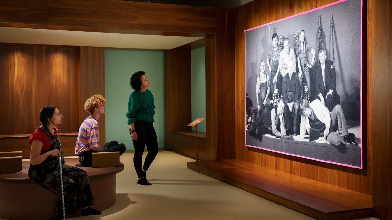 Photograph of a gallery exhibition space showing large tapestry hung on a wooden wall. The tapestry is made up of a black and white, archive photograph, from the early 20th century showing a group of white men and women gathered in a group holding museum artefacts in their hands or on their laps. They are surrounded by other museum exhibits and display cases. The tapestry is framed with a bright yellow border. Surrounding the tapestry is a wider wooden structure containing seating and alcoves. To the right of the image three gallery visitors are looking at the tapestry.