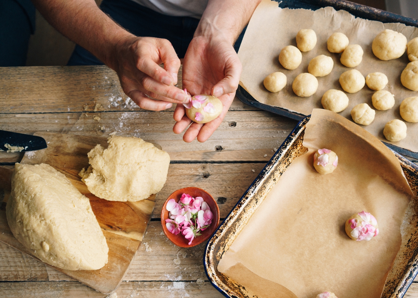 Photograph of a worn wooden kitchen table from above. A man's hands are decorating an uncooked dough ball with flower petals. On the tabletop is a wooden chopping board, two baking trays, lined with greaseproof paper and a small bowl containing flower heads and petals. In the baking trays are other uncooked dough balls.