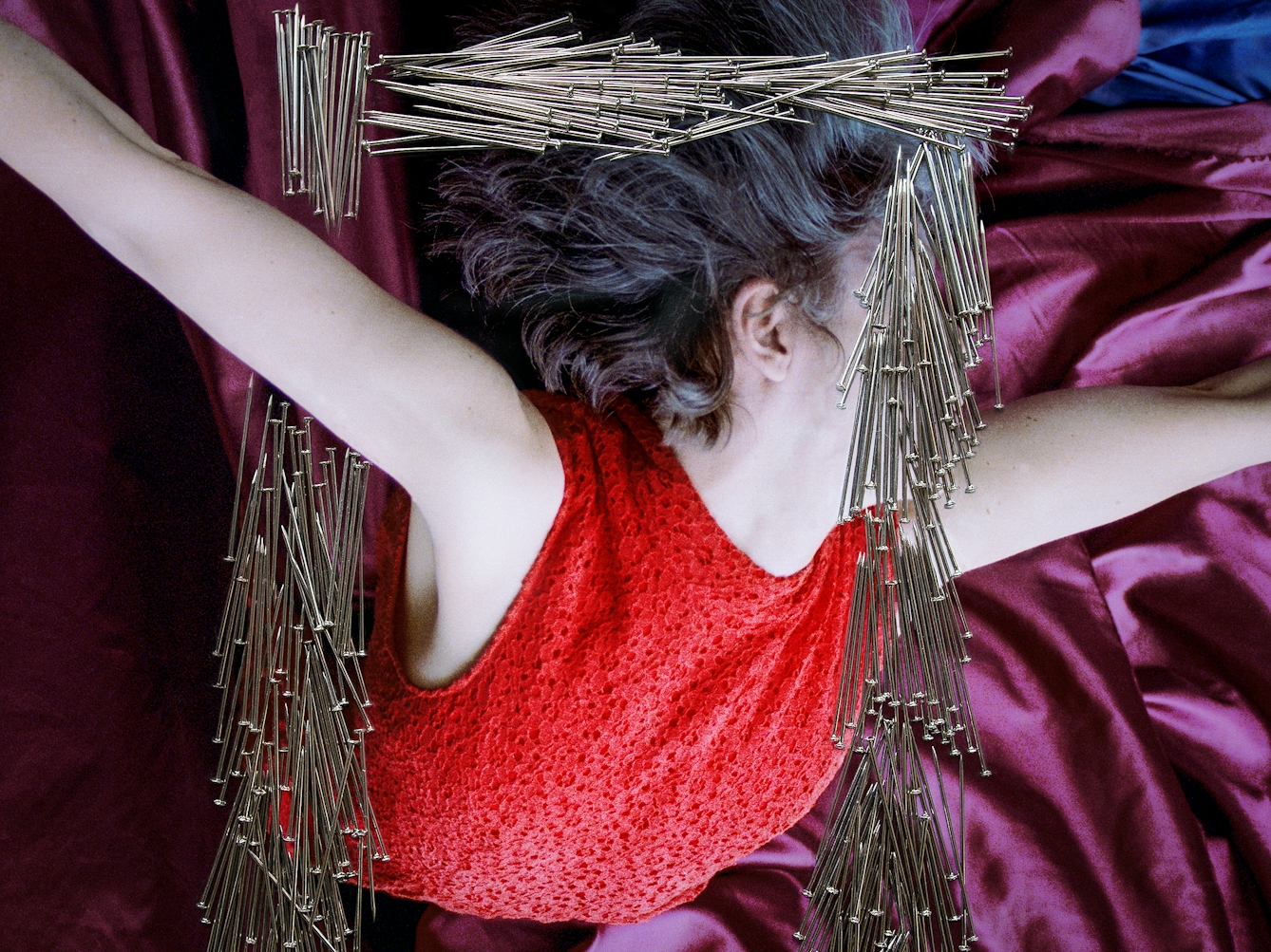 Detail from a larger artwork created with a colour photographic print of a female figure in a bright red dress, set against a purple and blue draped silk background. The figure is viewed as if from above her looking down. Her arms are extended out to the side. Her body is covered by groups of dress pins, laid on top of the photographic print. The pins are arranged in a square frame shape in the centre of the print. One side of this frame covers her face. Another side of the pin frame stops above and below her extended arm making it look as if she is bursting through the frame.