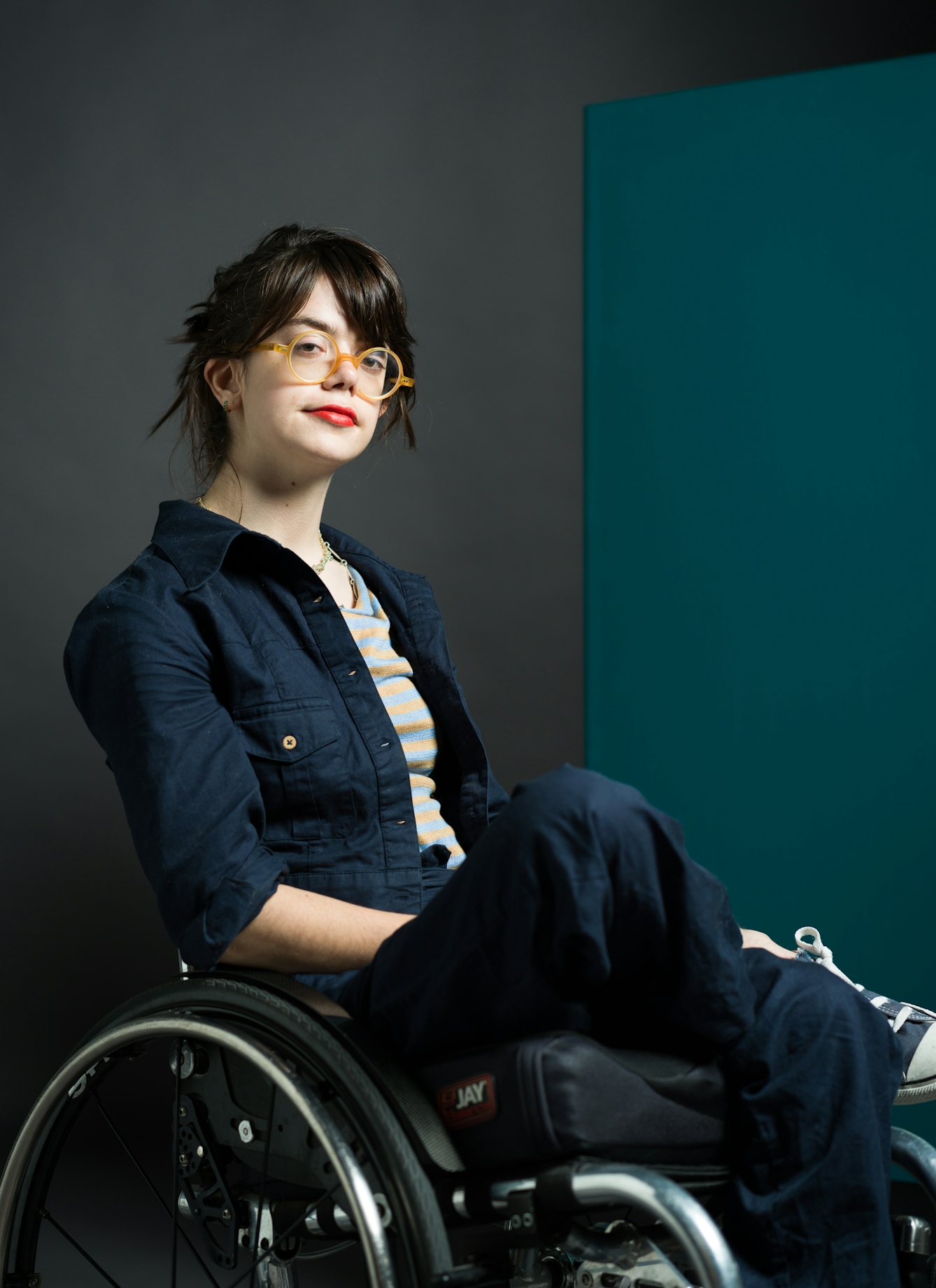 Colour photograph showing three-quarter portrait of Grace Spence Green, a white woman sitting in a wheelchair with one leg crossed over the other. Grace has dark hair, round orange glasses-frames, and bright lipstick. She wears dark denim overalls, a stripy t-shirt and trainers.