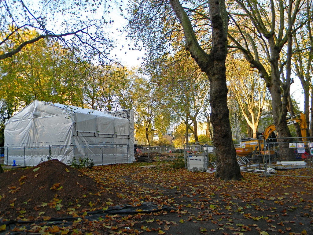 Photograph of St James's gardens in November 2017.  In the foreground is a mound of excavated earth.  Behind this is a high metal fence, a digger, and a construction site tent.