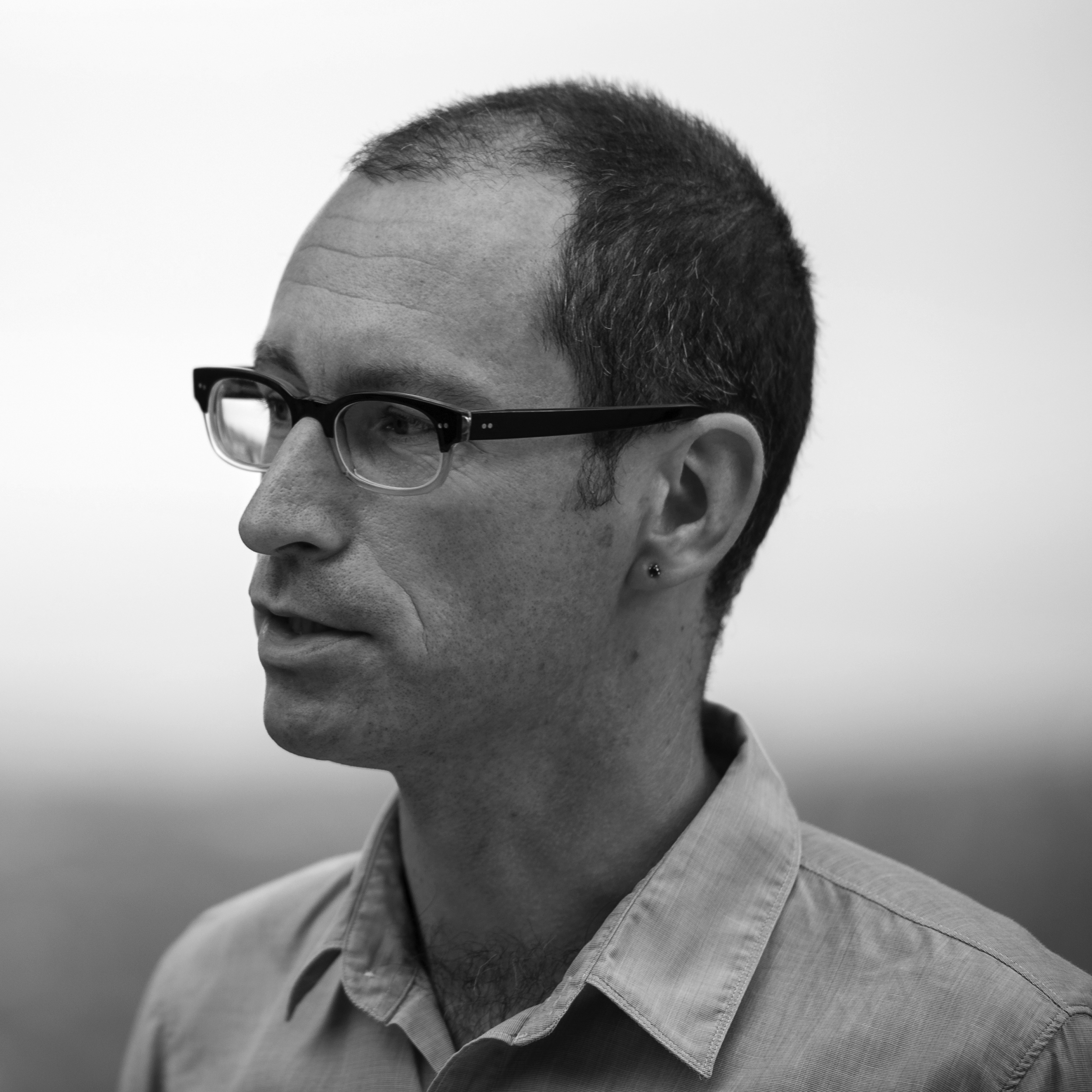 Photograph of the head and shoulders of a man in three quarters view, lips slightly parted as if in speech, an earring in his left earlobe, with short hair and wearing glasses.
