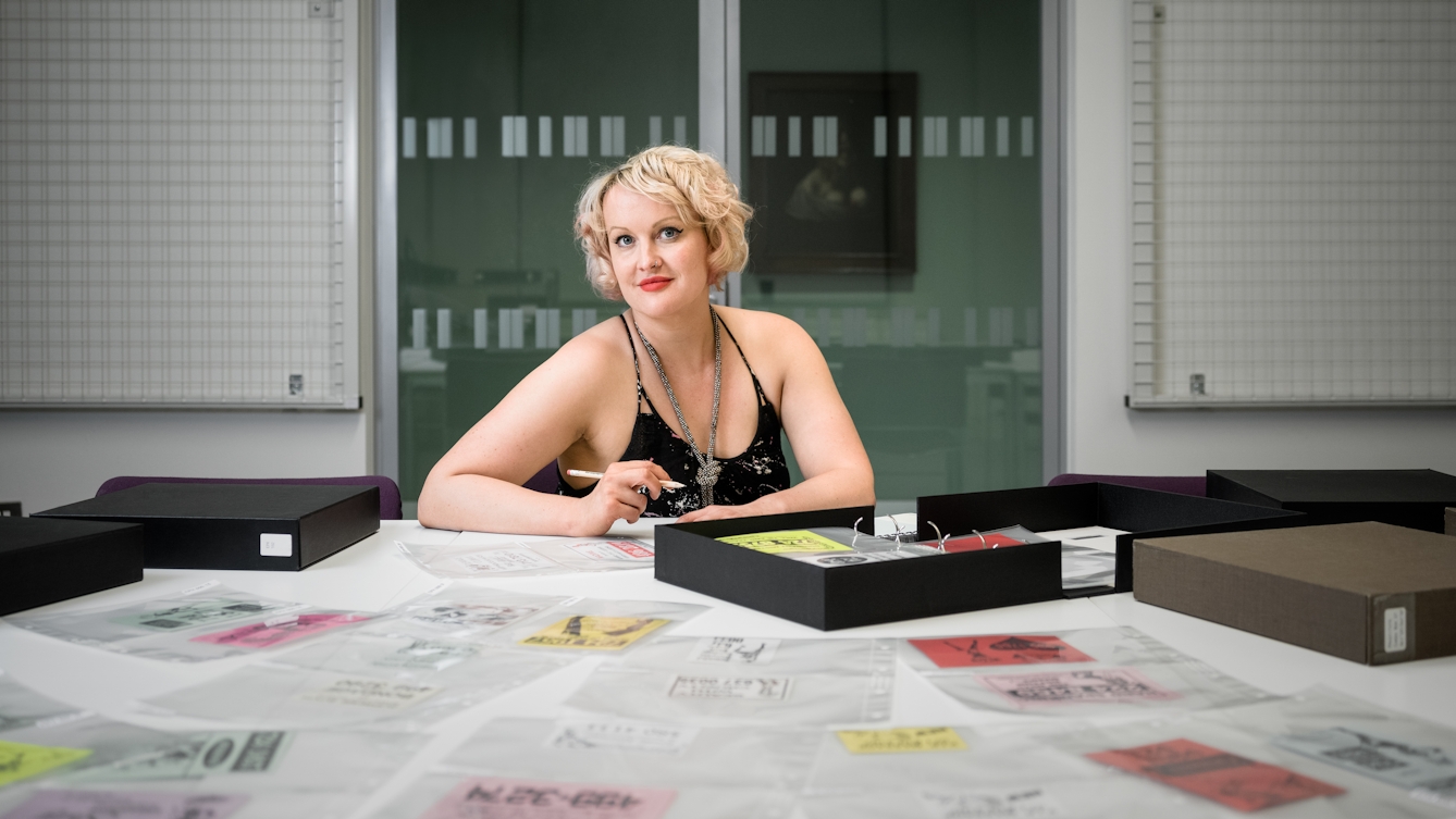 Photographic portrait of Dr Kate Lister sat at a table with a selection of archive material from the sex worker card collection at Wellcome Library laid out infront of her.