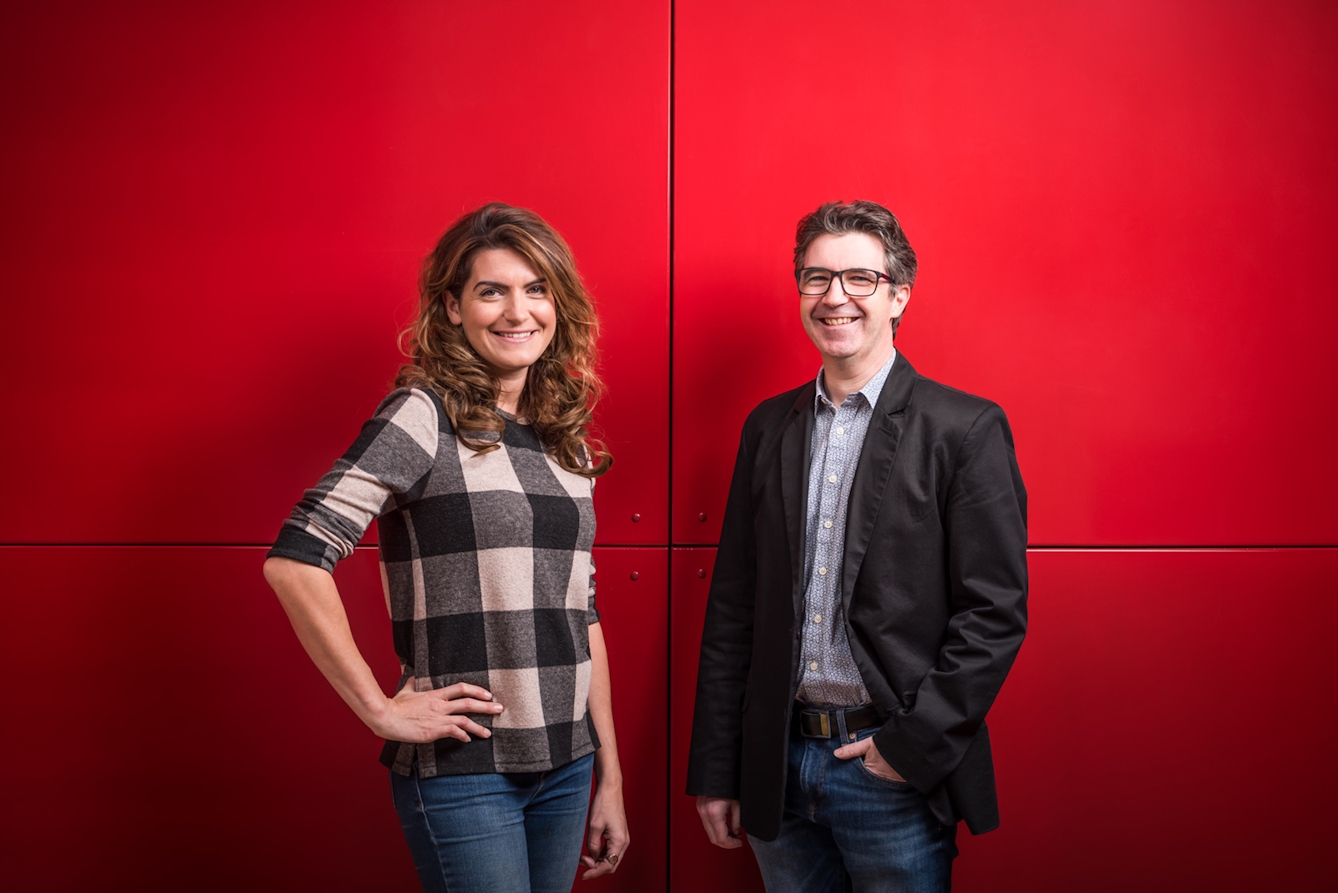 A portrait of a woman and a man standing against a bright red background.