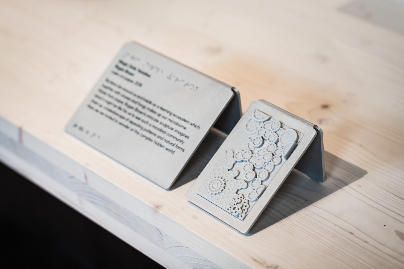 A photograph of a caption, black text shown as out of focus and displayed on a small silver display plaque positioned at 45 degrees to wooden surface, alongside a similar sized touch tile to allow visitors to feel a texture or shape of an exhibit without touching the object itself. The touch tile shows what looks like a series of cogs shown in relief.