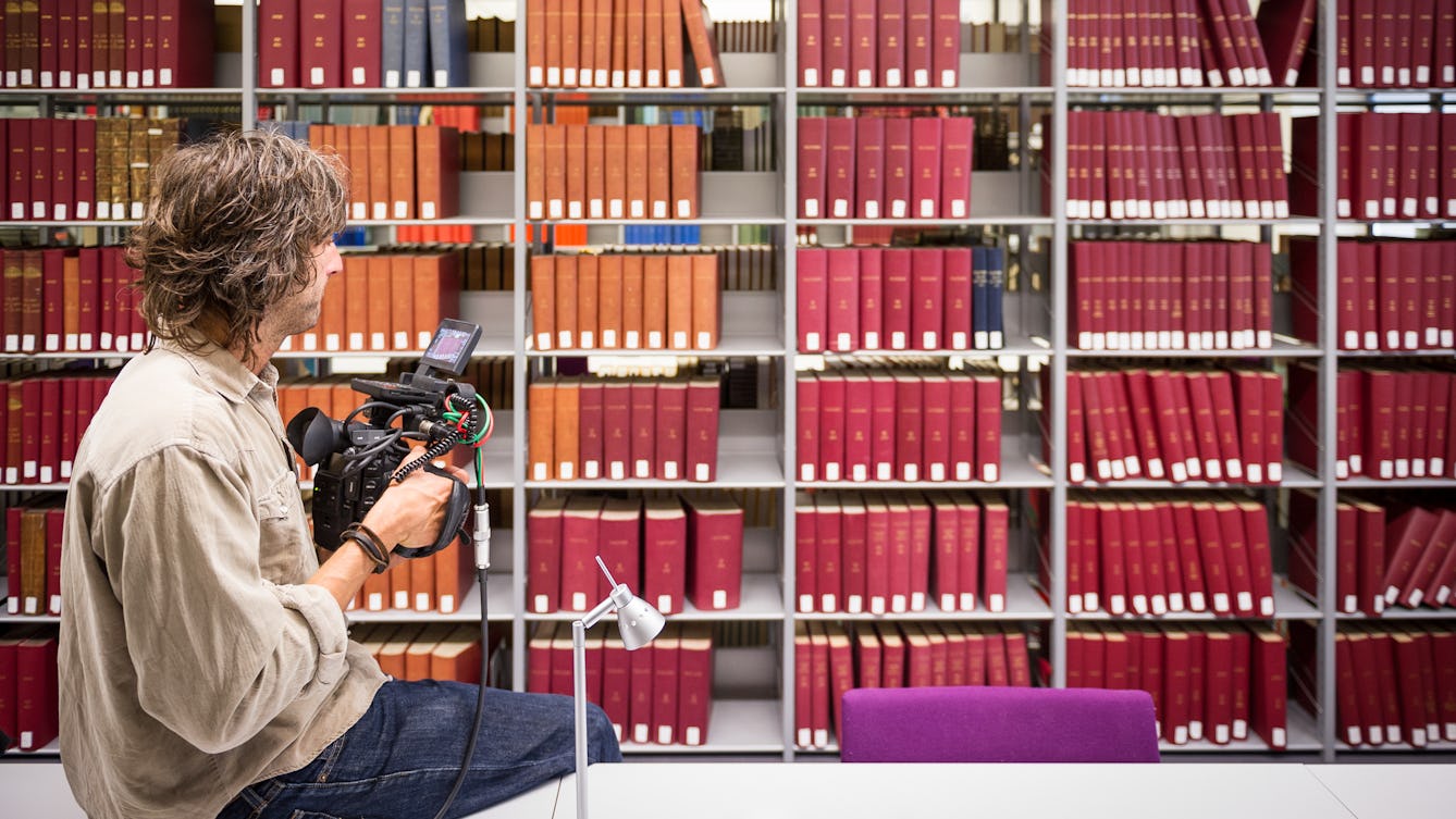 Photograph of a library bookshelf filled with books, spines facing outwards. All the spines are either red orange or green in colour. In the foreground is a man perched on a long white tabletop holding a large professional film camera with a small screen and wires protruding. He is looking towards the bookshelf.