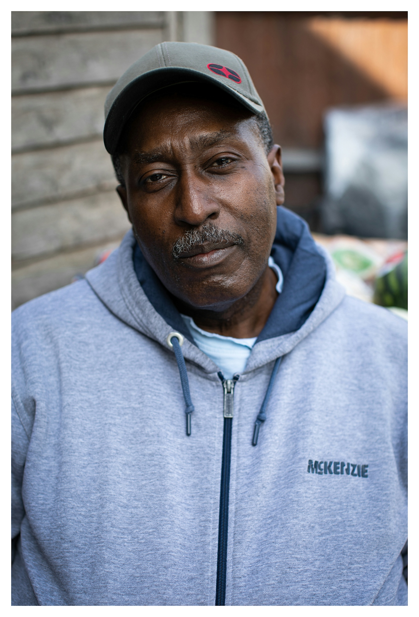 Photographic portrait of a Black man with a greying moustache, wearing a baseball cap and a light blue hooded top. He is looking to camera with his head slight tilted to the left. He has a kind, open expression on his face. Behind him are out-of-focus elements of a garden scene.