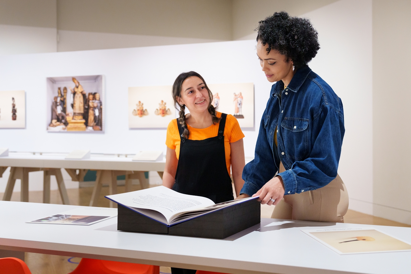 Photograph of a gallery exhibition space showing large photographic prints mounted on the walls and long tables containing prints and books. Two people are stood at one of tables looking at a large book sitting on book rests. They are looking at the book and chatting. The colours of the room are light whites and yellows.