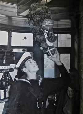 Photograph of Alan Beckett, a Royal Engineer, placing a hot cross bun onto a stack of buns. He is looking up at the hot cross buns and smiling. There is a woman sitting below him looking down. He is wearing a Navy uniform. 