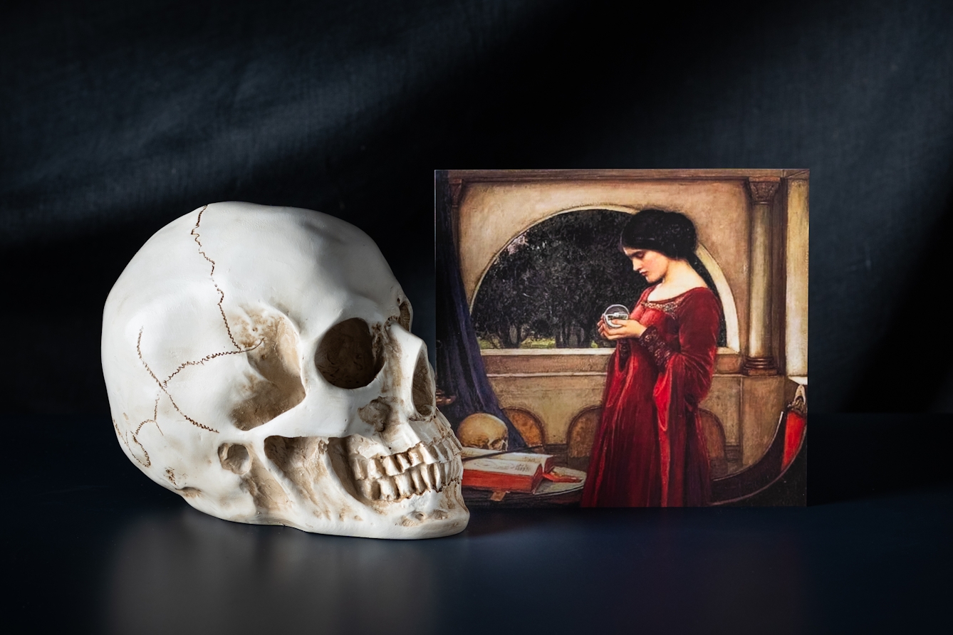 A still life photograph showing a reproduction of a section of a painting depicting a renaissance women. She is wearing a long red dress and is holding a crystal ball in her hand. She is standing in front of an arched window with dark draped curtains. On the table beside her is a large book with red edged pages and a human skull. A human skull is also presented next to  the painting The scene is presented against a dark cloth background.