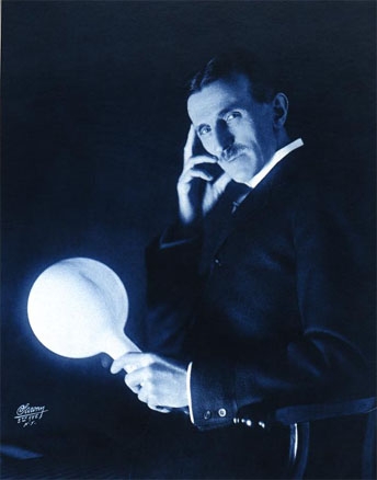 Black and white photograph of Nikola Tesla sitting with one hand on his face and the other grasping a bulb that is glowing with light.