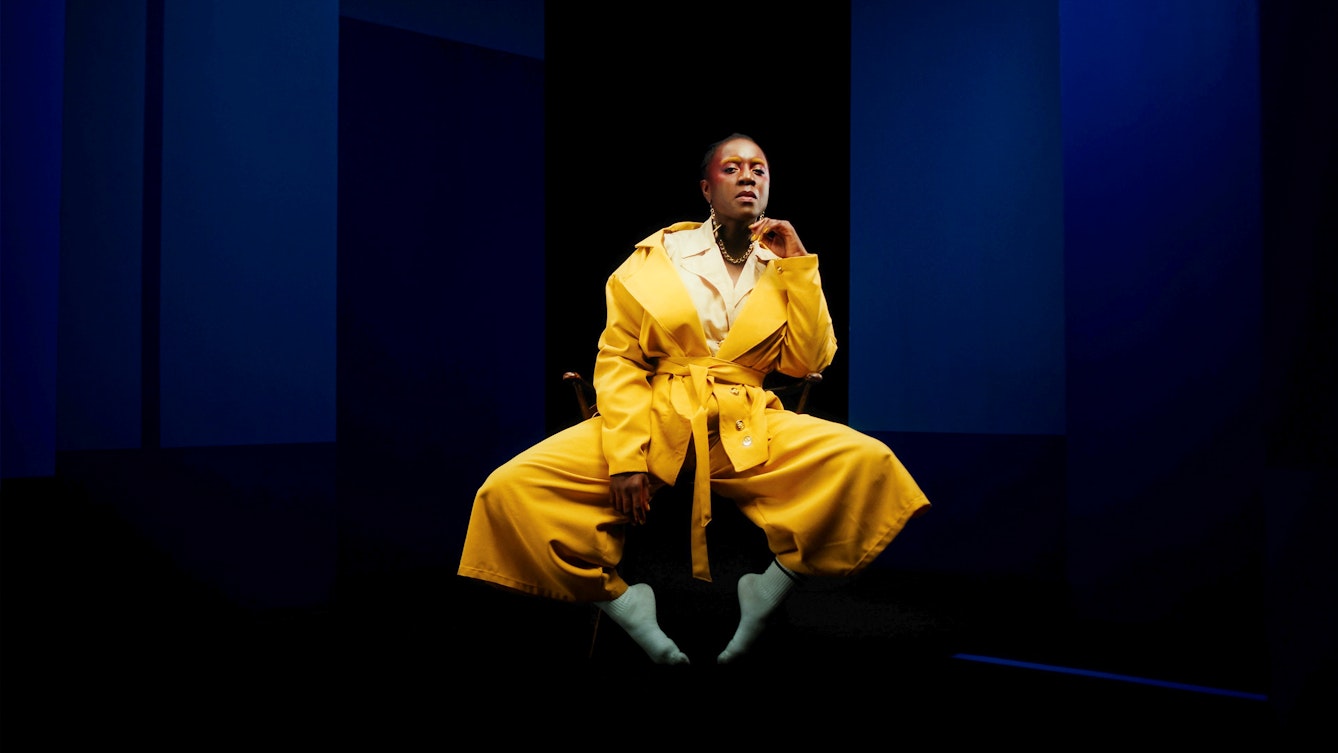 A still from a colour film. The image shows an individual in bright yellow trousers and jacket, sat on a chair with their left elbow resting on the arm rest, hand raised up by their face. They are looking to camera with a questioning tilt and expression to their face. Behind them is a black and blue background. They look as if they may be sat on a stage of some kind.