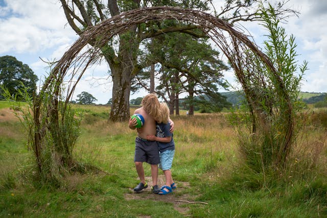 Photograph of two children standing in a field, underneath an arch formed out of trees. The taller child is holding a green and blue football and is wearing grey shorts and blue Crocs. The other child is wearing a blue t-shirt, light blue shorts and sandals. They both have blonde hair.

There are some trees behind them and a blue cloudy sky.