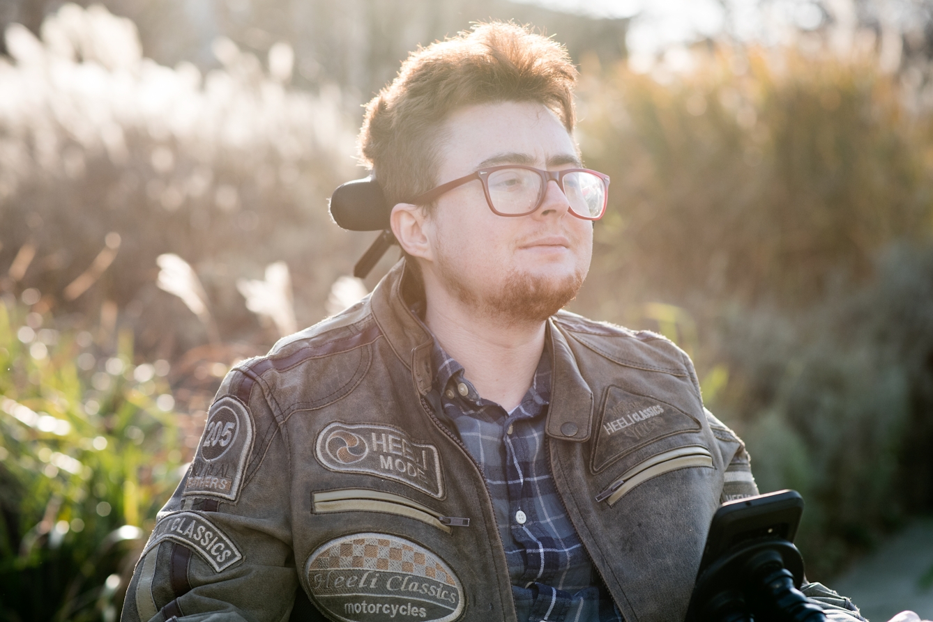 Photographic portrait of a young person looking into the distance off to camera right. Behind them are long grasses and shrubs lit from behind by a low sun.