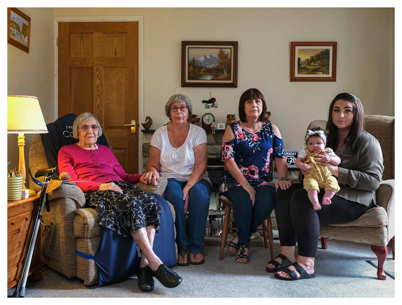 Family group photograph showing five generations of the same family. The great-great-grandmother sits on the left in a large armchair, by her side sits the great-grandmother who is holding her hand. Next to her sits the grandmother who rests her hand on the knee of the great-granddaughter who is holding the great-great-granddaughter on her knee. They are all seated on chairs in a living room, surrounded by framed pictures and furniture. They all look straight into the camera.