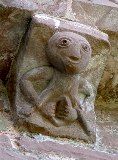 Colour photograph of a stone carving showing a grotesque figure with a smiling face and two hands holding open its vagina.
