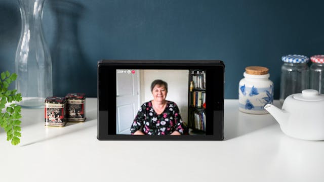 Photograph of a tablet on a white desk. The tablet screen shows a photographic portrait of Dr Teresa Hillier. On the desk there are a number of objects including a teapot, jam-jars, a plant and a vase. The wall behind the desk is painted dark blue. 