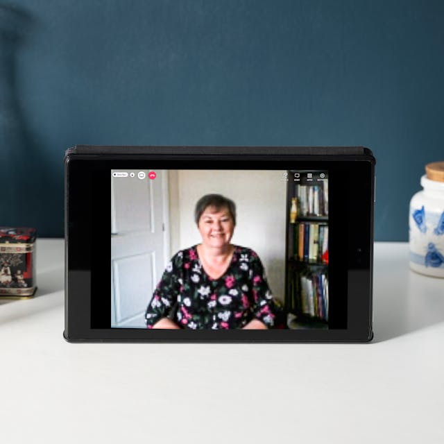 Photograph of a tablet on a white desk. The tablet screen shows a photographic portrait of Dr Teresa Hillier. On the desk there are a number of objects including a teapot, jam-jars, a plant and a vase. The wall behind the desk is painted dark blue. 