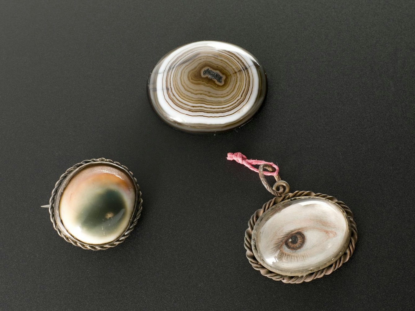 Photograph of 3 amulets placed on a textured grey background. All are amulets against the evil eye. One depicts a drawing of a human eye, the other 2 display more abstract patterns of concentric rings and colour patches.
