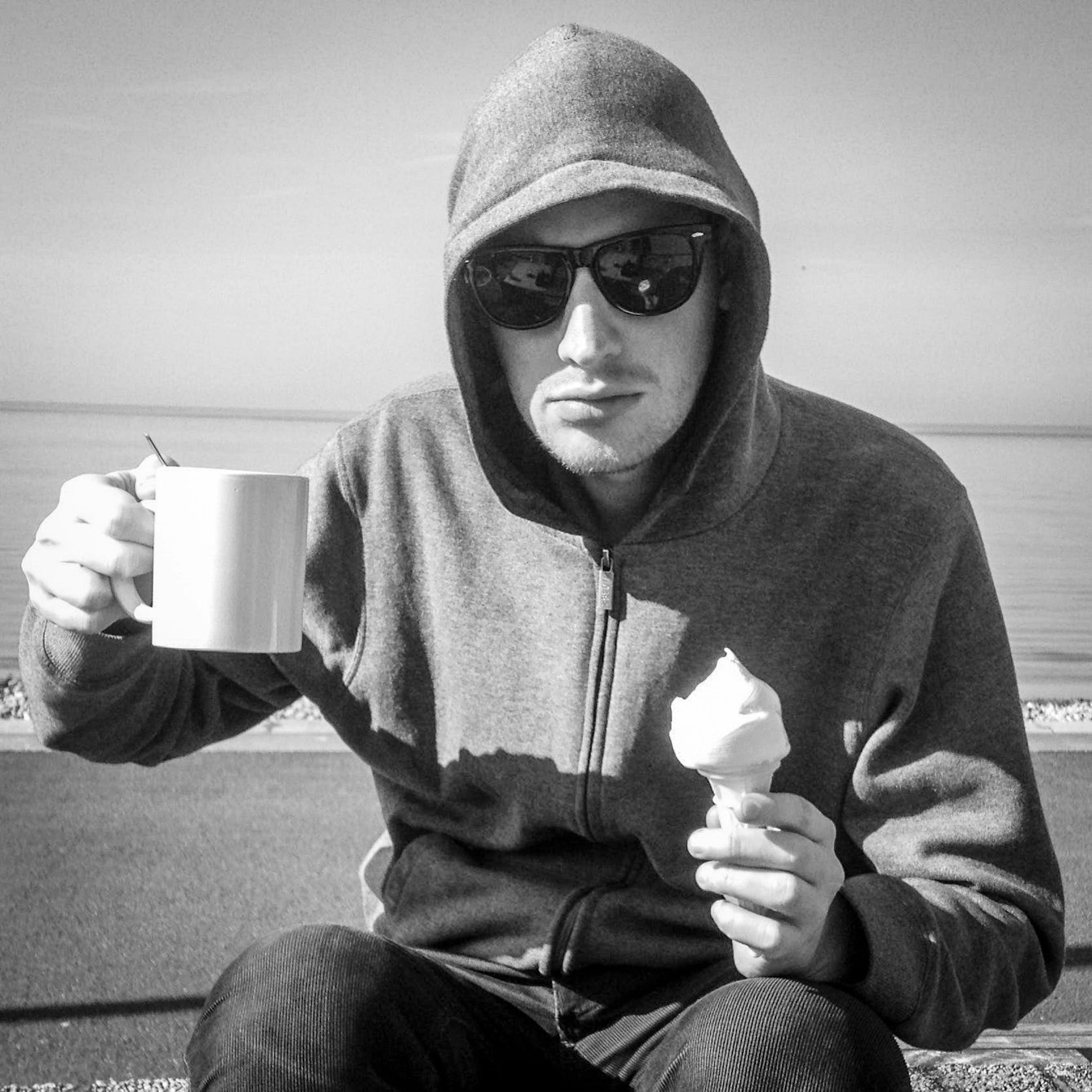 Black and white portrait of Rob Bidder sitting by the sea holding an ice cream cone in one hand and a white mug of tea in the other. He has the hood of his jumper over his head and is wearing a pair of sunglasses.
