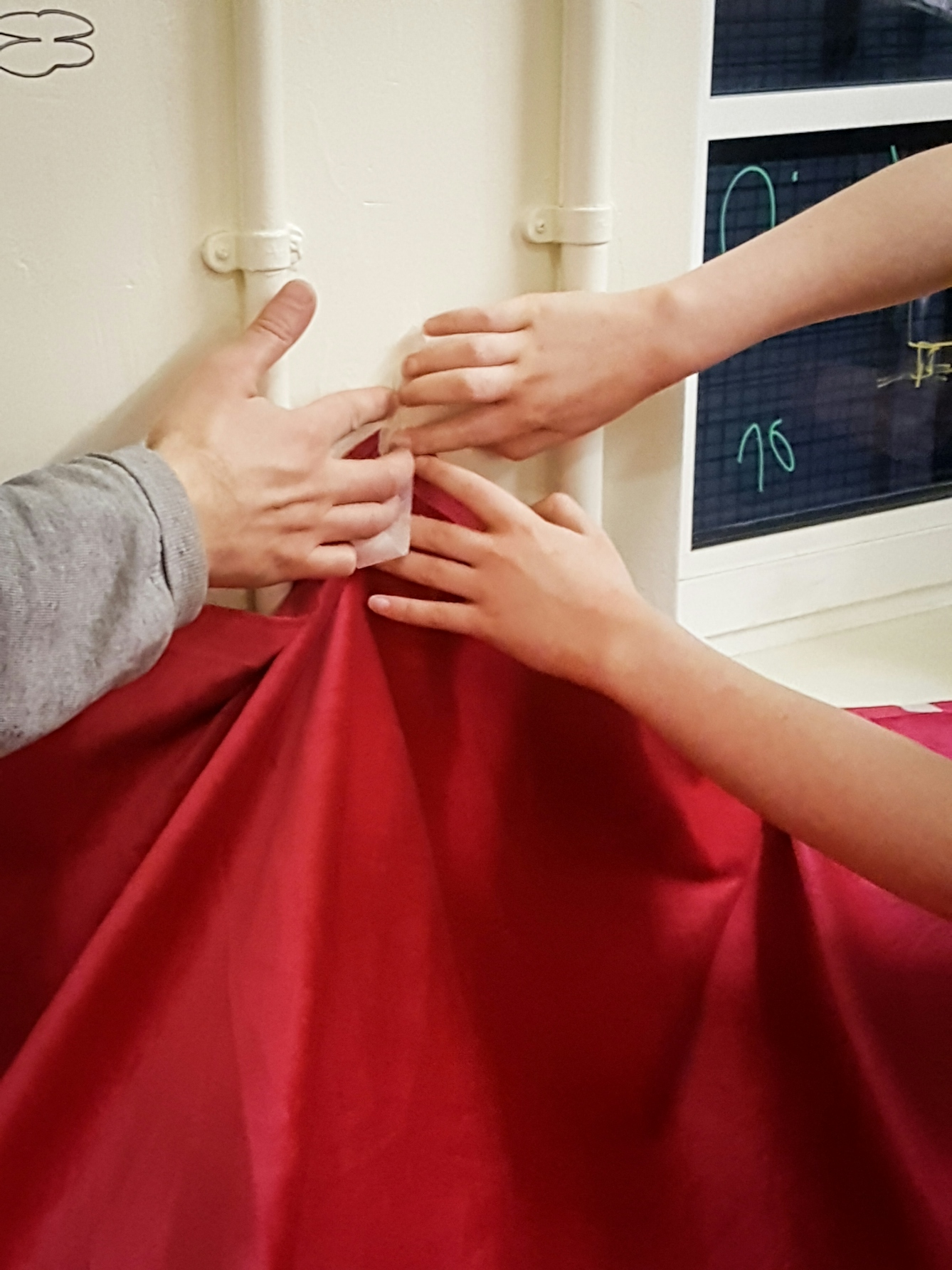 A photograph in portrait orientation showing a close up of a large piece of red fabric being held up against a magnolia painted wall by three hands, one of which belongs to a young person, the others a child. Parallel to one another are two pipes painted magnolia running up the wall, and in the top right of the image the bottom corner of a blacked out window can be seen. 