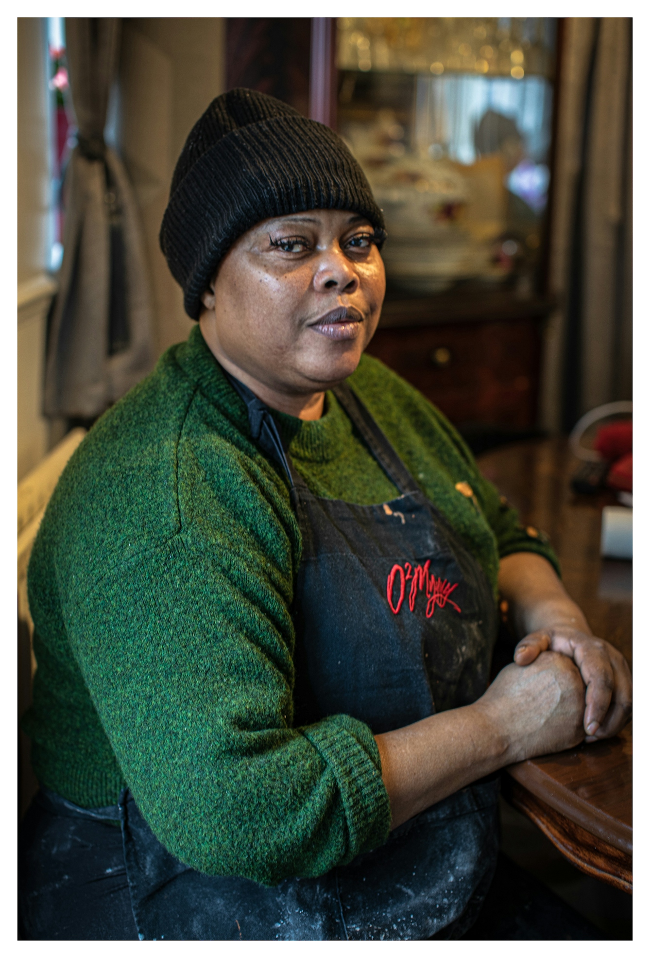Photographic portrait of a Black woman seating at a table indoors. She is wearing a green knitted jumper, a black wooly hat and a cooking apron. Her hands are gently clasped together on the tabletop. She is looking straight to camera with a strong, quiet but confident expression.