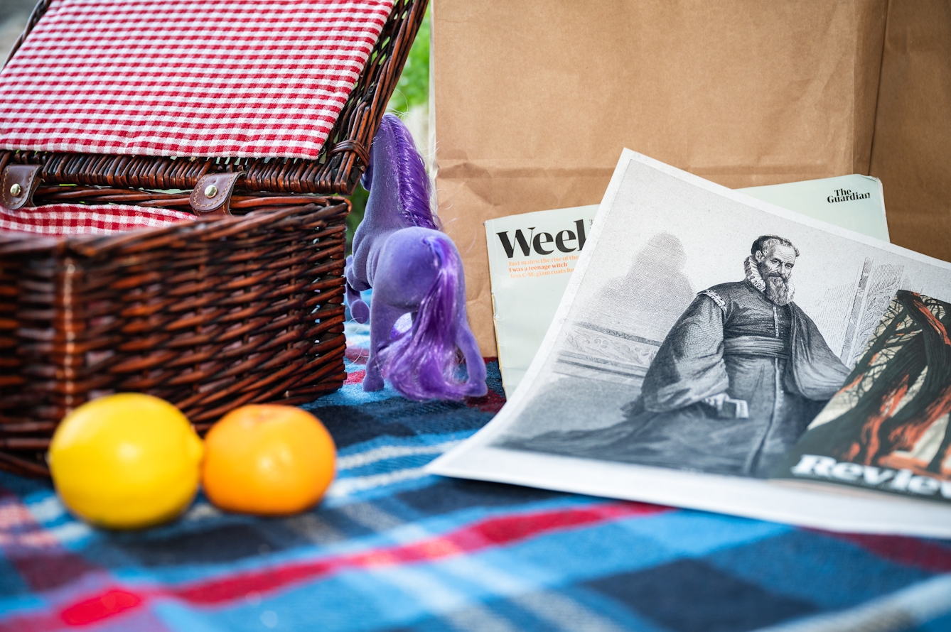 Photograph of a checkered picnic blanket, fairly closeup. On the left is an open wicker picnic hamper, in front of which is a lemon and a clementine. To the right of the image is a collection of newspaper supplements within which is a black and white print of a bearded man wearing long robes and a ruff. In the centre of the image is the rear end of a purple unicorn disappearing behind the picnic hamper.