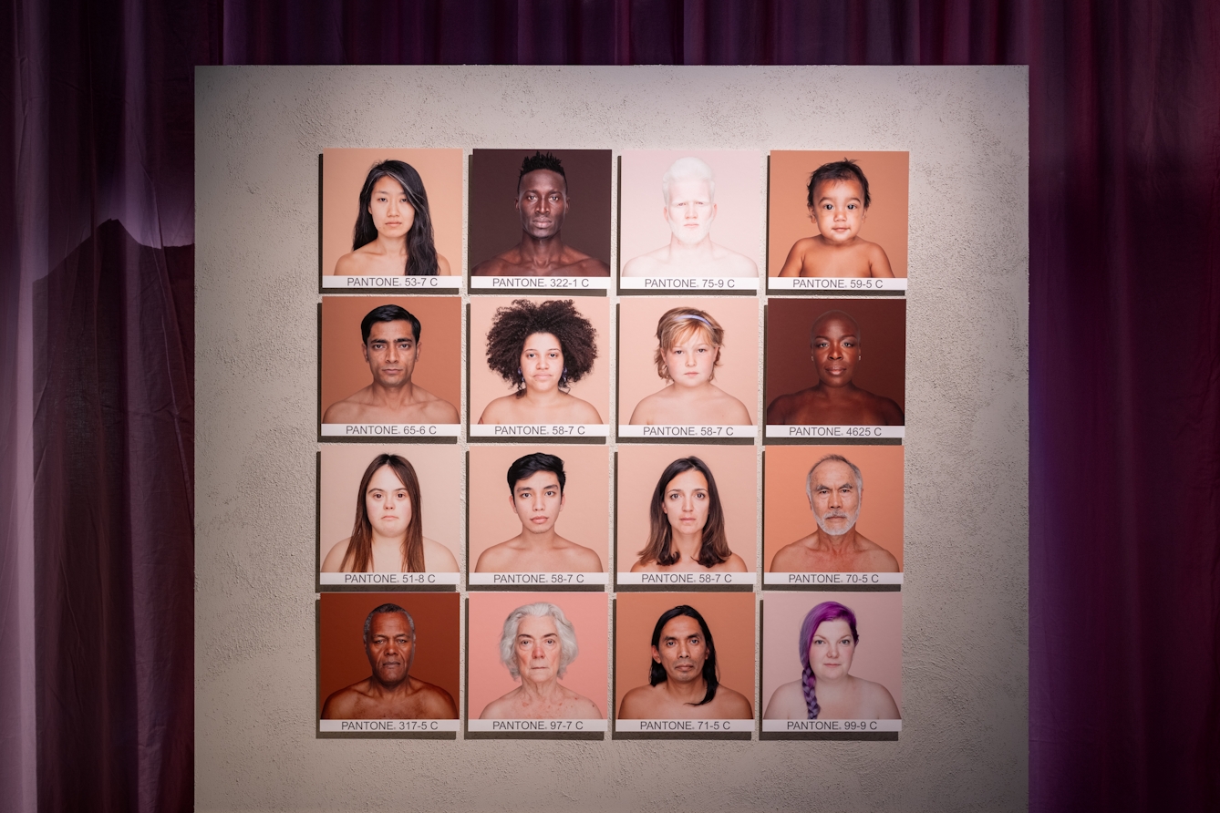 Photograph of a grid of 4 by 4 square portraits of 16 people's head and shoulders. The background colour for each portrait matches the hues of the person's skin. Under each image is a Pantone reference. 