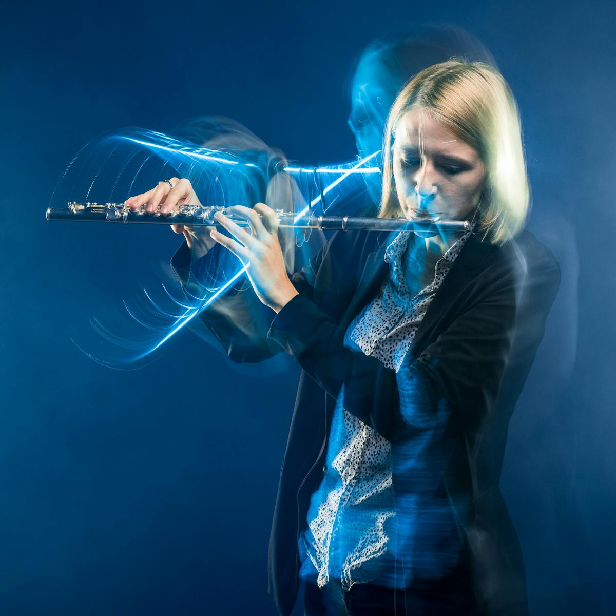 Photograph of a young woman playing a flute against a blue background. The slow exposure of the camera causes her movements to be traced across the image as a blur. She is surrounded by wisps of smoke.
