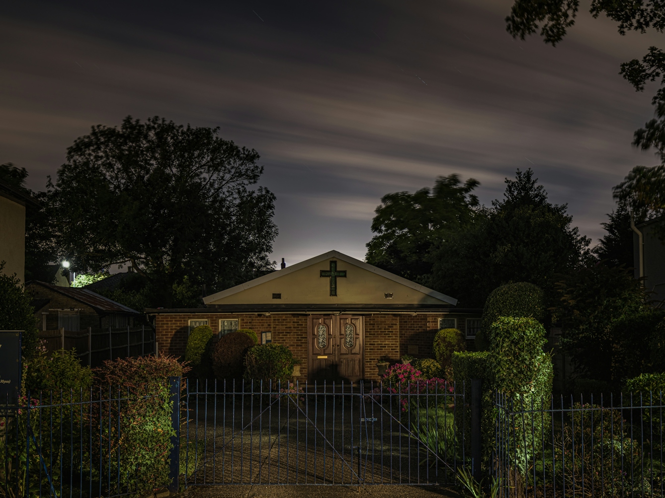 Photograph of Light on the Hill Spiritual Sanctuary, Dartford, captured at night.  The building has a pitched roof with a cross at the apex and doublers wooden doors.  The building is detached and surrounded by foliage, with a blue metal gate in the foreground.