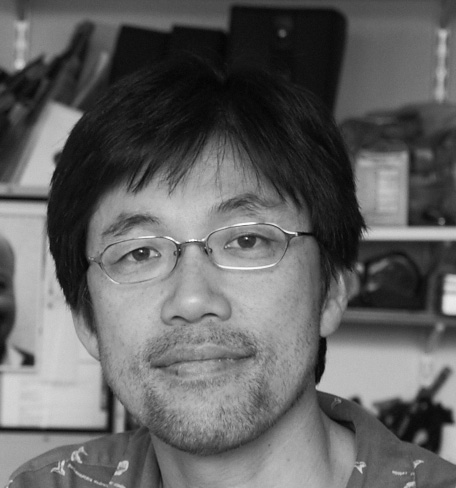 Black and white photograph of a Japanese man wearing small wire-framed glasses and looking straight at the camera. The background is blurred.