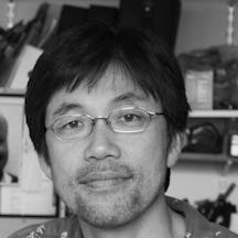 Black and white photograph of a Japanese man wearing small wire-framed glasses and looking straight at the camera. The background is blurred.