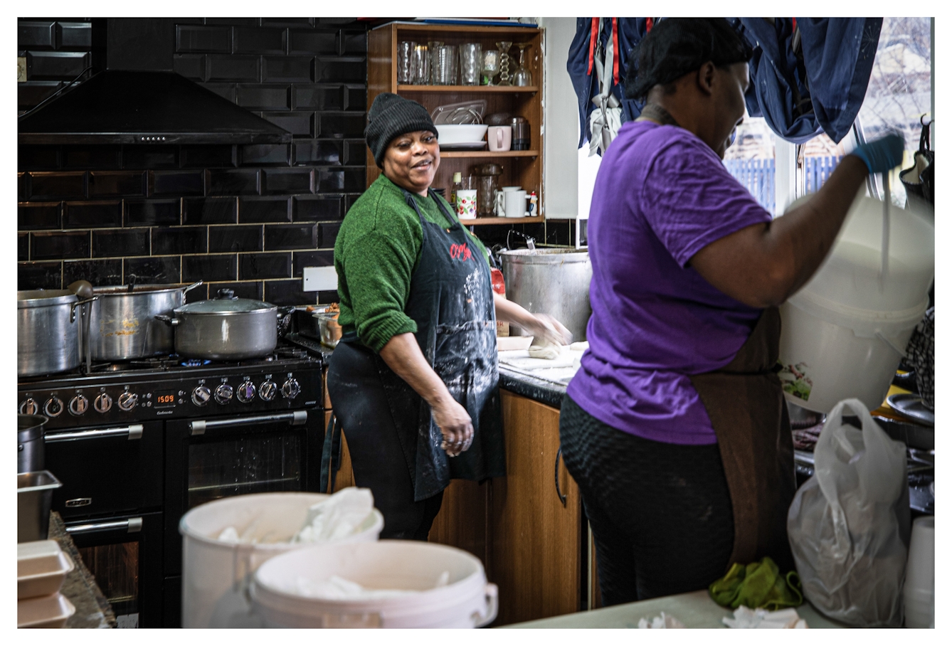 Photograph of a kitchen scene where cooking on a large scale is taking place. The stove is covered in large pots and pans. Two Black women are working in the kitchen, smiling and joking as they work.