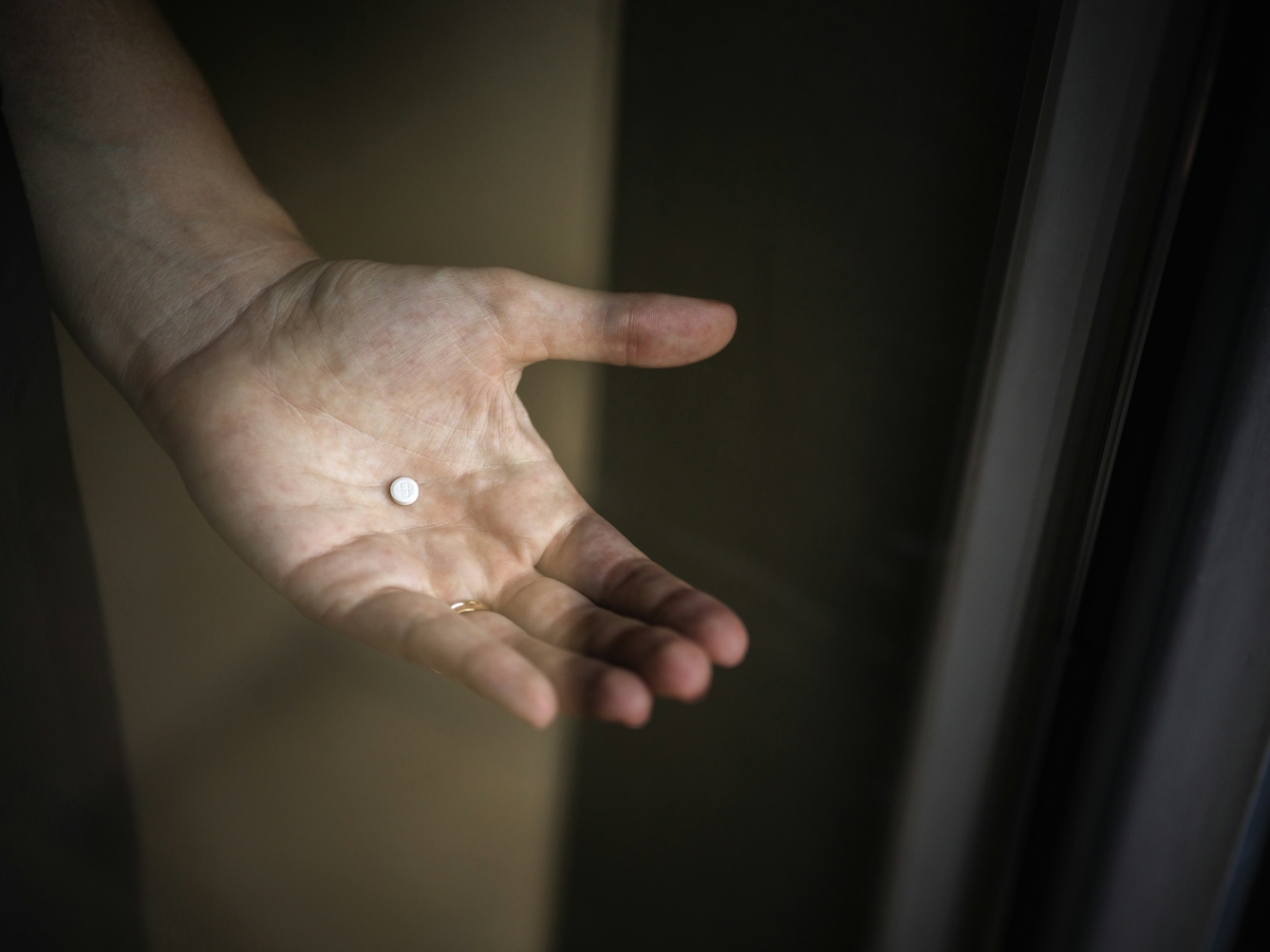 Photograph of a woman’s hand holding an immunosuppressant prescription tablet, captured through a window.  The marking on the tablet looks like the letter G in lower case or the number 9.  There is a slight reflection in the window.