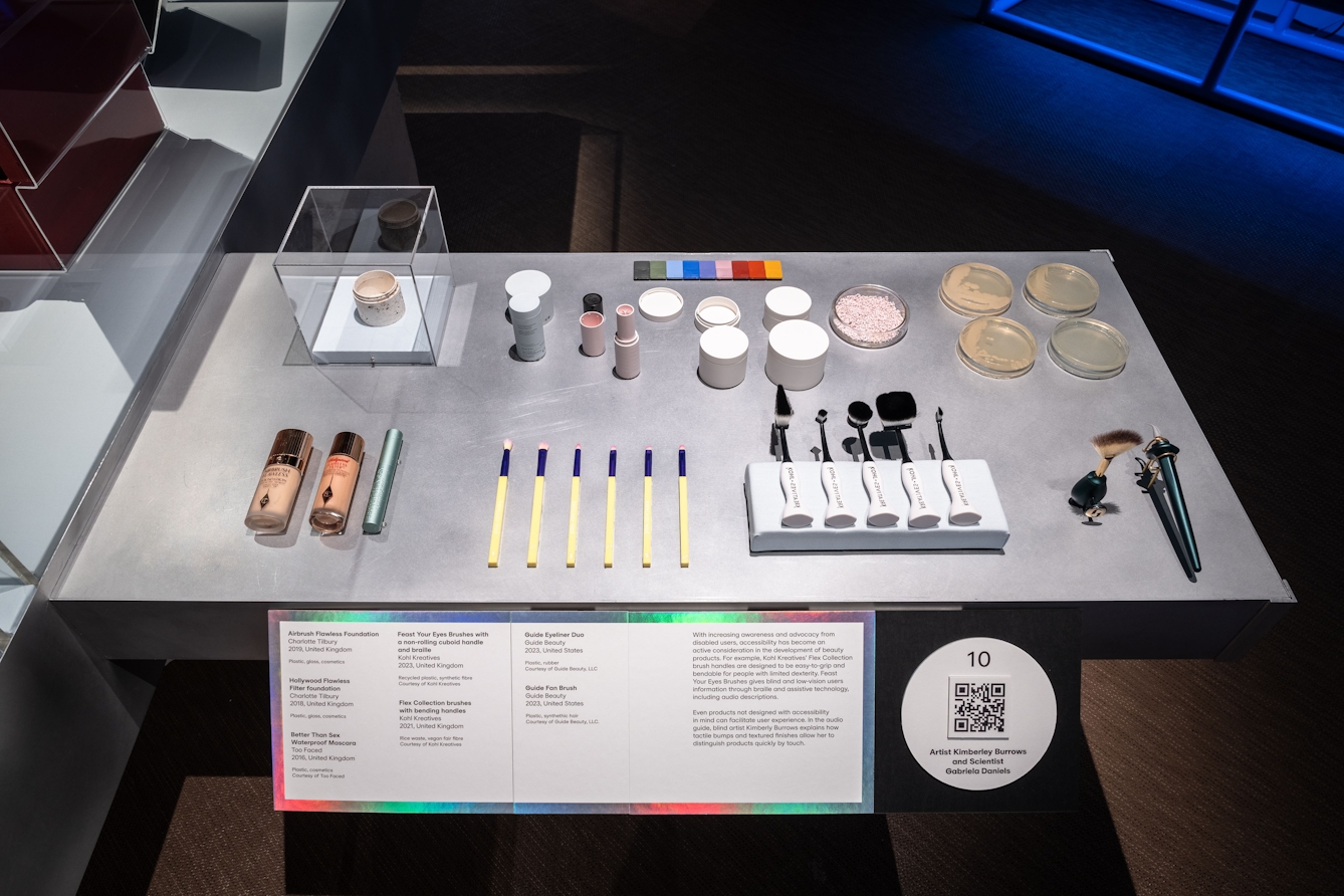 A table-top in an exhibition space with a caption board in front of it. On the table are instruments and containers used for make-up and beauty products. Sime are designed for accessibility with larger handles and brushes.