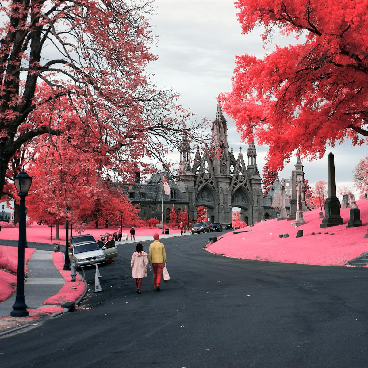 Infrared photograph of the entrance to Green-Wood Cemetery. In the bottom left portion of the image two people are walking towards the Neo-Gothic entrance to Green Wood Cemetary. The road has a series of parked cars of which one has a door open. To the right of the frame there is a large tree towering over several grave stones. The pink hues replacing the greens of the grass and trees are a result of the infrared technique.