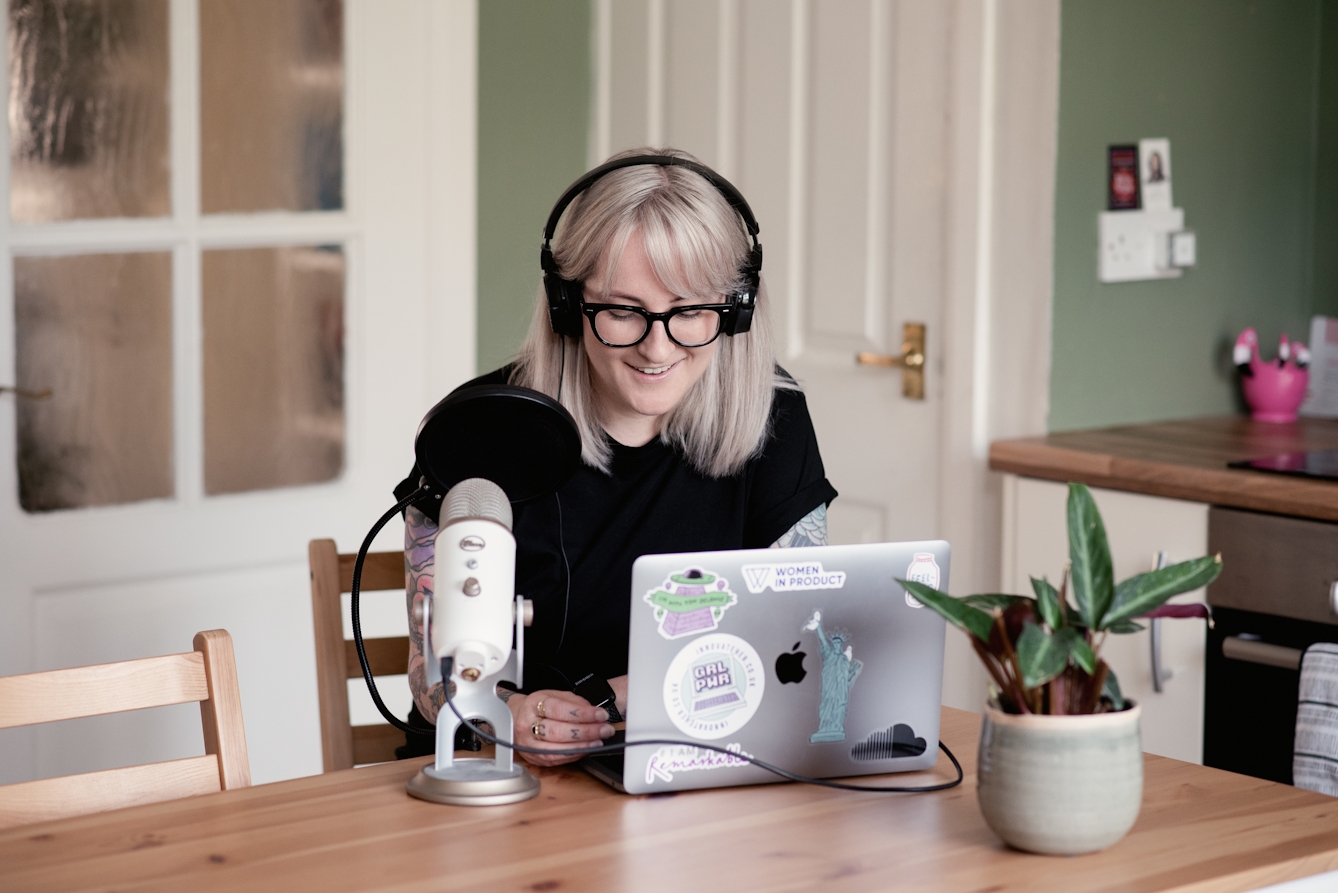 Photograph of a young woman with shoulder length blond hair seated indoors in a home environment at a kitchen table. She is wearing black glasses and a pair of large headphones over her ears. She has on a black t-shirt. She is smiling and looking down at the laptop which is resting on the tabletop in front of her. To her right is a professional looking microphone in a stand on the table. Her laptop is covered in stickers. Also on the table is a small house plant. In the background are the doors and countertop of the surrounding kitchen.
