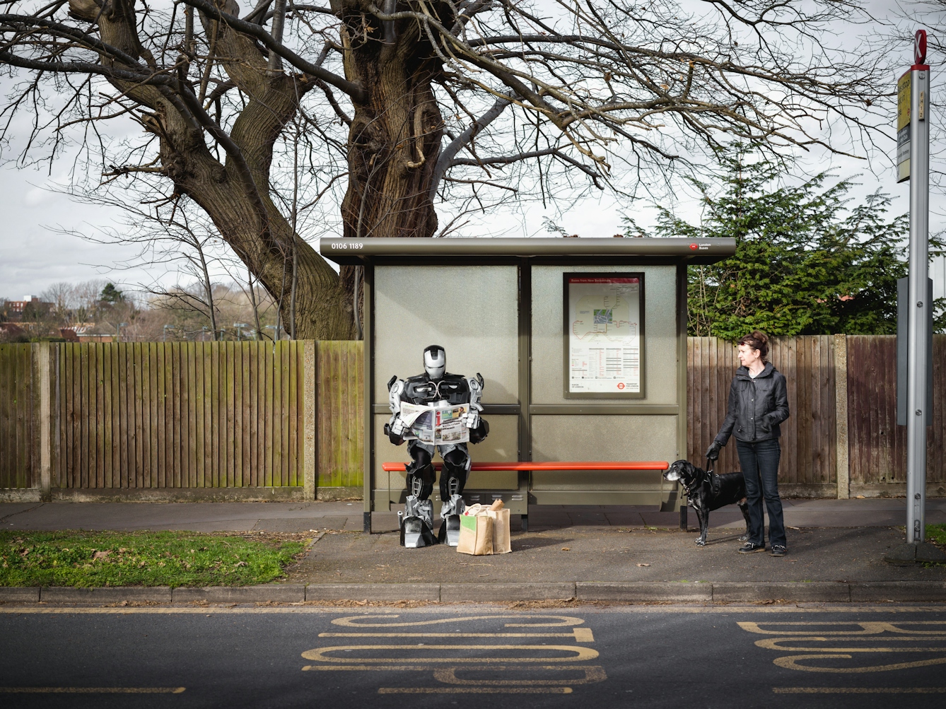 Photograph of a large robot sat at a bus stop reading the paper. At its feet are shopping bags. A woman with a dog is also waiting at the bus stop.