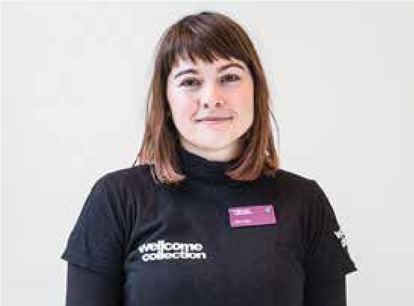 A young woman with shoulder-length brown hair is smiling and looking straight at the viewer. She is dressed in black with a Wellcome Collection logo on her t-shirt.