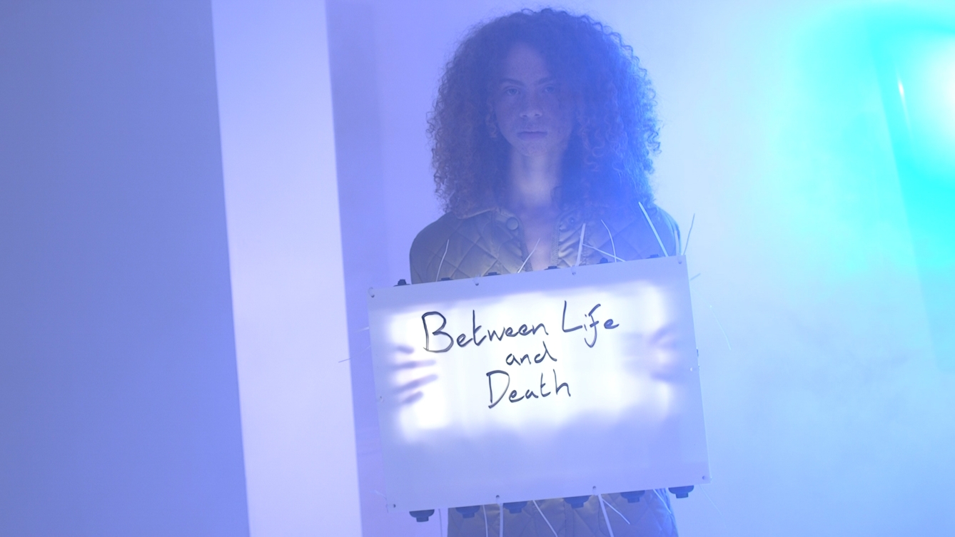 Colour film still of a woman from the waist up holding an illuminated sign with the words, 'Between Life and Death' written on it. The general hues of the image are purples and blues.