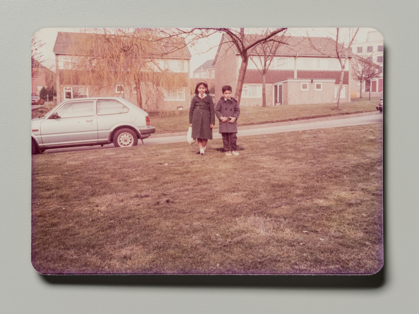 Photograph of a family photographic print resting on a grey background. The photo shows a young girl and boy in school uniform, standing on a grassy lawn in front of a 1960 housing estate.