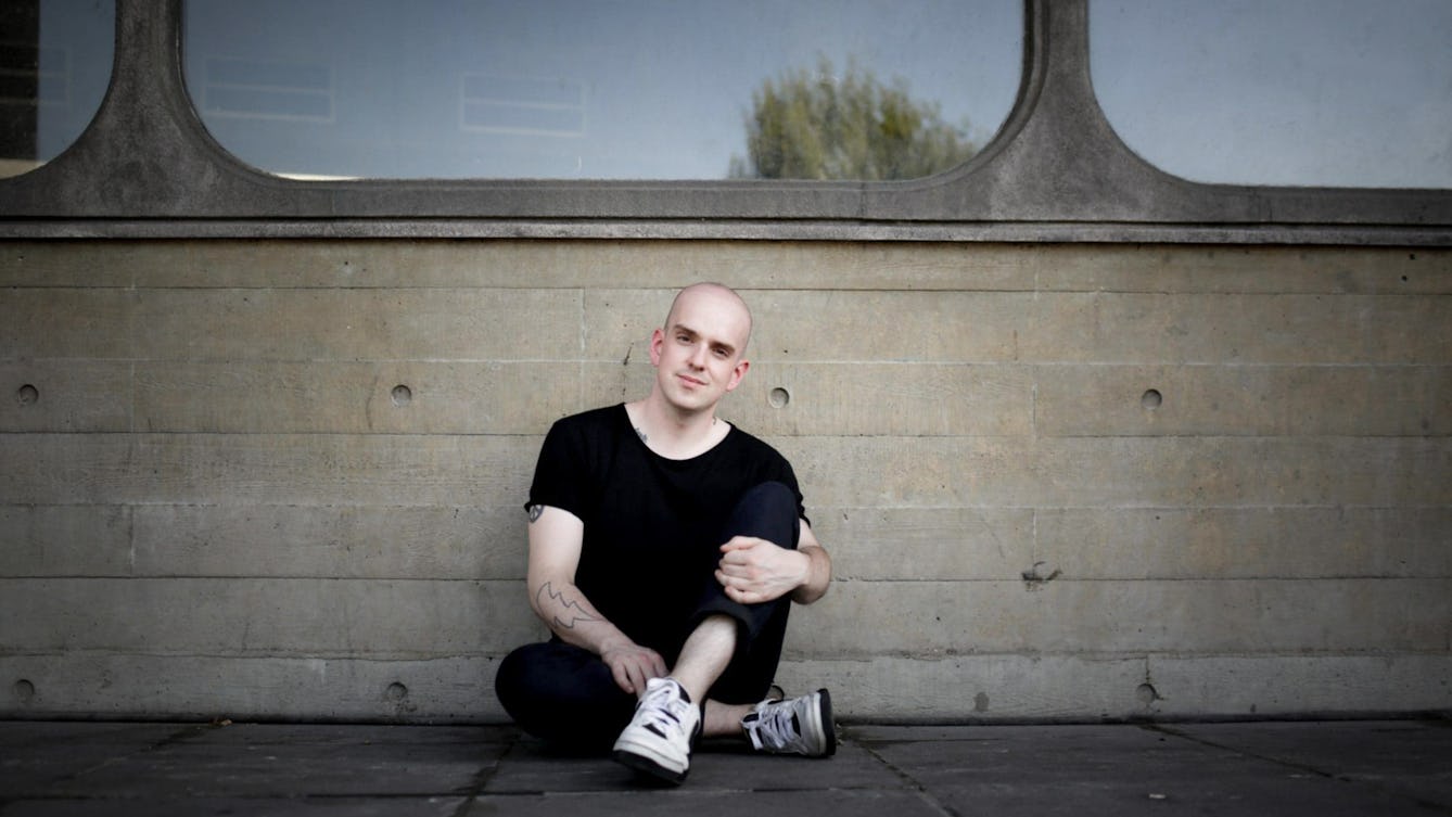 Photograph of a man dressed in black trousers and t-shirt, sitting on the pavement against a concrete wall containing large glass windows. 