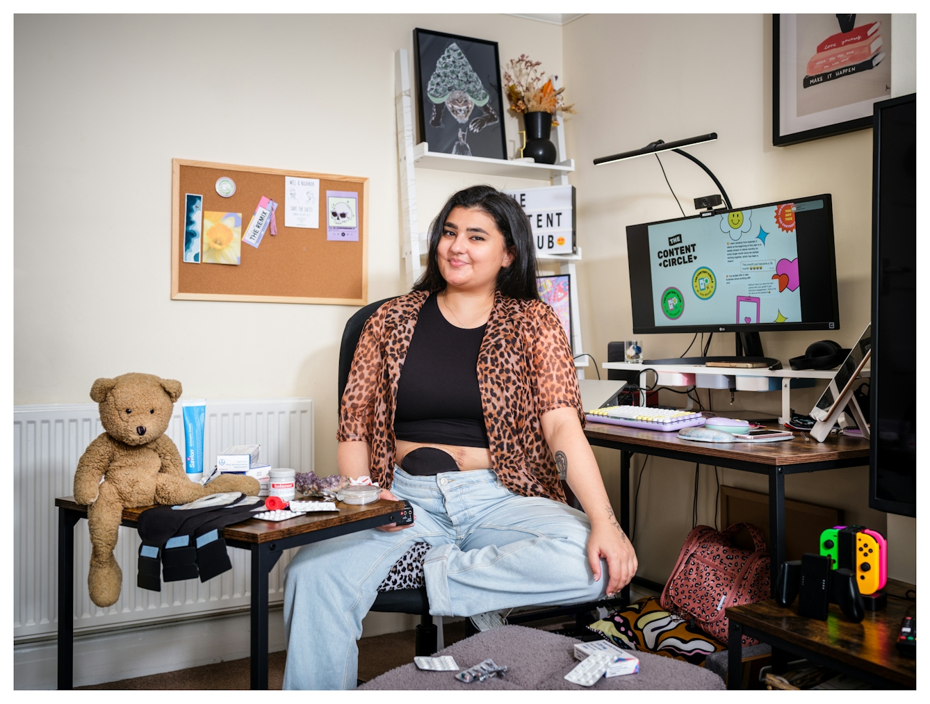 Assisted Self-Portrait of a woman sat on a swivel chair in her home office. She is smiling to camera, surrounded by medicines and her black stoma bag is visible above the waist band of her jeans. On the table to the left a brown teddy bear sits gazing at the camera, with his leg dangling off the edge. In her right hand the remote trigger she used to capture the image is just visible.