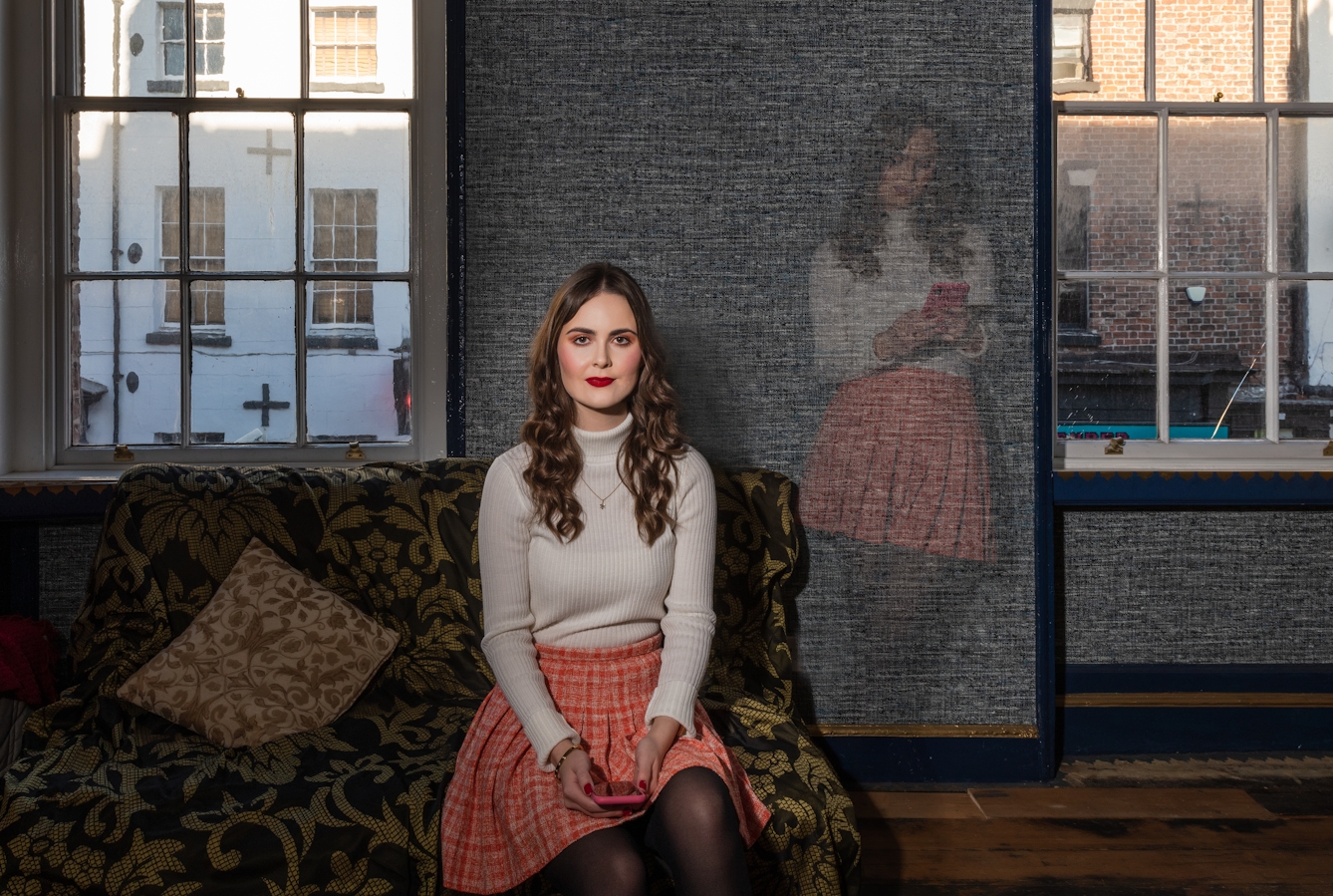 A portrait of Emily Bashforth sitting on a patterned sofa. She is wearing a cream jumper, orange plaid skirt, red lipstick and is holding a pink iPhone. She has long brown wavy hair and is looking directly at the camera.

Behind the sofa are two windows, showing buildings outside. Between the windows, there is a panel on the wall where there is an image of Emily superimposed. She is standing up and looking at her phone. 