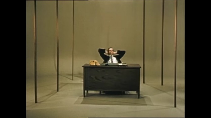 Still image from film of man sitting at a desk with his hands behind his head looking relaxed