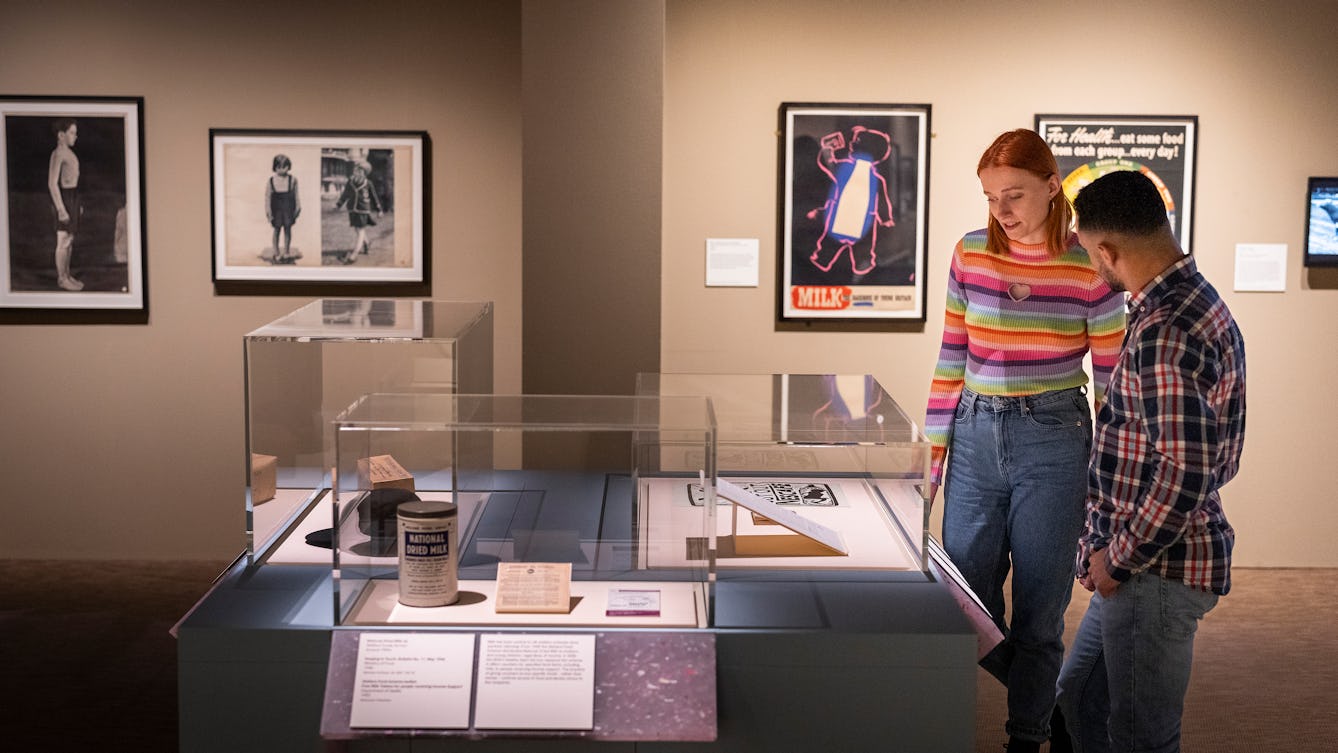 Two exhibition visitors looking at objects in an exhibition space. The visitors are looking at print materials and objects displayed in a glass box. Behind them are framed works hung on the wall.