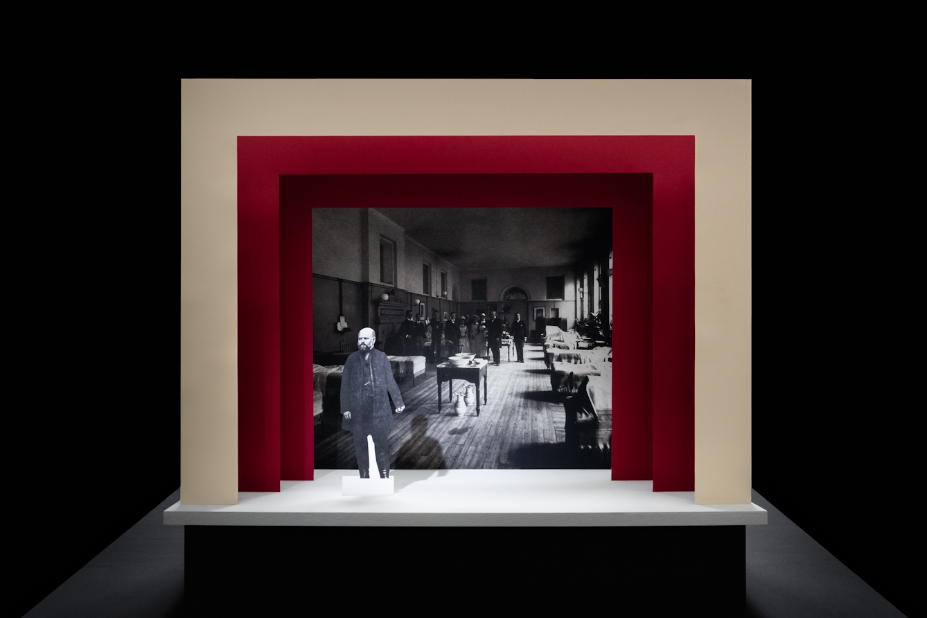 Photograph of a simple theatre stage set, made out of card. The background surrounding the stage is black. The stage floor is white and the framing of the stage is made out of 3 square edged arches, each one smaller than the other, receding backwards. The first arch is cream coloured and the other two are a red. On the stage is a small cut out photograph of a man with a beard from the early 20th century. Behind him forming the backdrop is a black and white archive photograph of a maternity ward, also from the early 20th century.