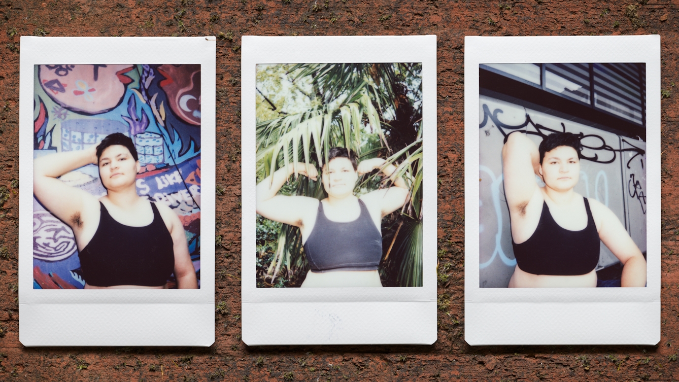 Photograph of three Instax Mini instant film prints in a line, resting on a textured brick surface. All three prints show the same man. The print on the right shows the man wearing a black cropped vest standing against a graffitied wall with his right arm raised up behind his head to show his armpit hair. The print in the centre shows him nestled within the leaves and branches of a tree. He is raising both arms to the side, elbows bent, revealing his armpit hair. The print on the left shows him again standing against a colourful graffitied wall with his right arm raised up behind his head to show his armpit hair.